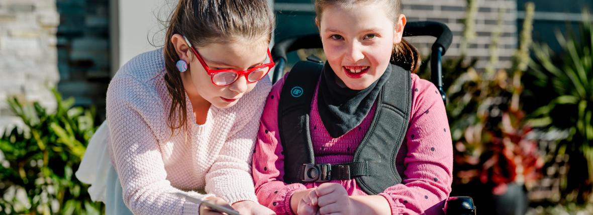 Inclusive outcome - Equitable access to health services for Australians, when and where they need them. Two young girls both dressed in pink and with glasses, leaning in together, smiling girl facing camera with special needs in wheelchair