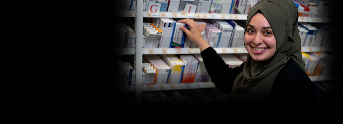 National Digital Health Strategy partners - Young woman wearing olive green hijab and black top, in a pharmaceutical setting reaching behind her to select medications