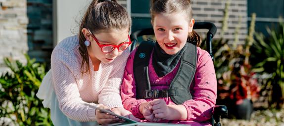 Inclusive outcome - Equitable access to health services for Australians, when and where they need them. Two young girls both dressed in pink and with glasses, leaning in together, smiling girl facing camera with special needs in wheelchair