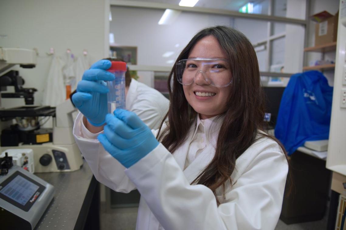 Person in a lab coat and blue gloves holding a clear tube with a red cap in a laboratory.