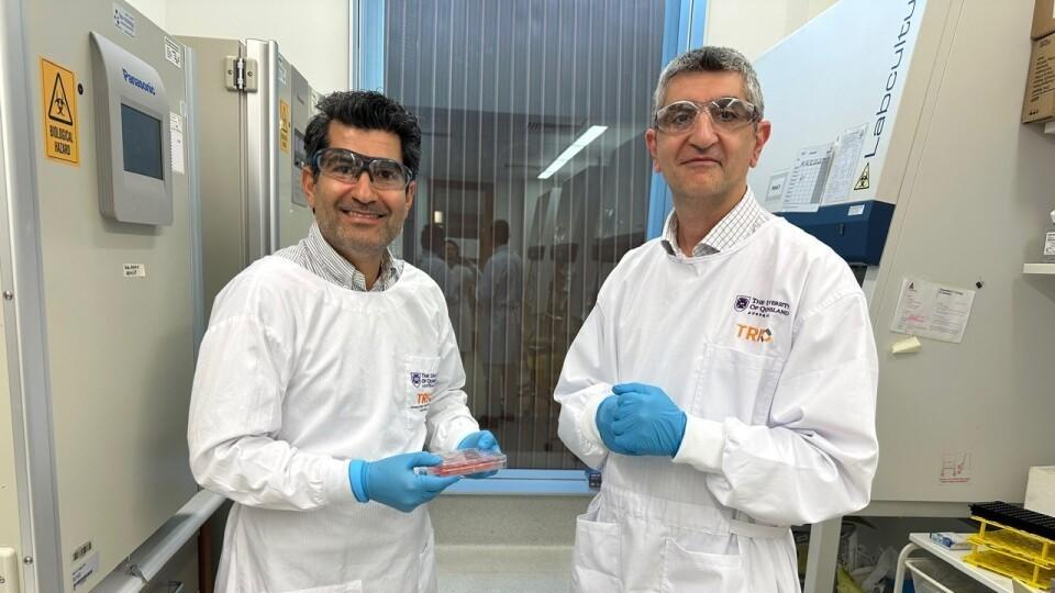 Two people in white lab coats, safety glasses and blue gloves standing in a laboratory, one person is holding small container.