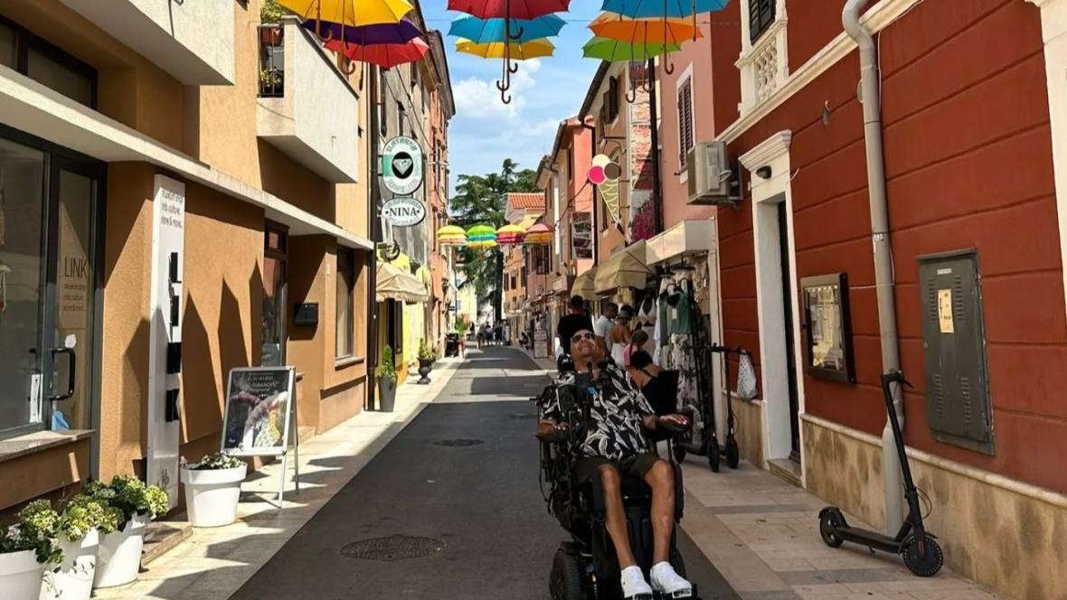 Colorful umbrellas suspended above a narrow pedestrian street in a European-style town, with people walking, a person in a motorized wheelchair, and a parked electric scooter along the vibrant, café-lined path.